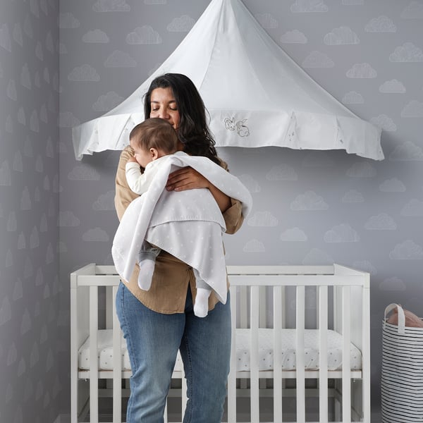 Person holds baby by crib with cloud wallpaper and canopy. Baby swaddled in recycled polyester blanket.