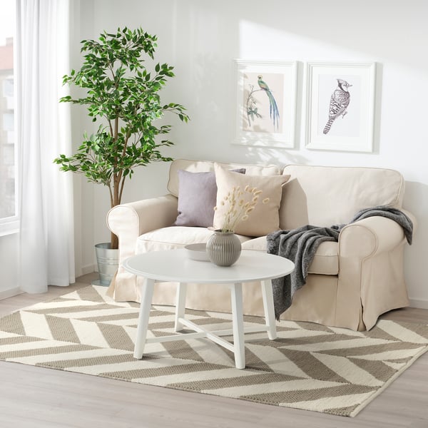 The LOKSTALL rug, beige & grey striped, wool & cotton, under a white couch, near a round white table, with a large plant beside.