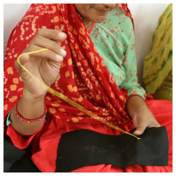 A person in a red and green patterned outfit is carefully sewing a yellow thread into a small black pouch.