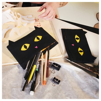 Black cat-shaped pouch with yellow eyes and colourful nose, holding pens. IKEA MÄVINN, 100% cotton, hand on zip.