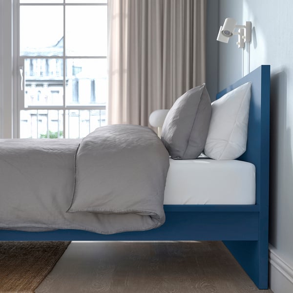 Modern minimalist blue MALM bed against window, taller than floor rug, with neutral linens and pillows, wall lamp nearby.