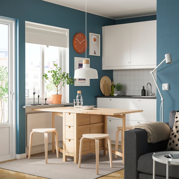 Wooden drop-leaf table with drawers, stools. Modern kitchen with teal walls, white cabinets, hanging lamp.