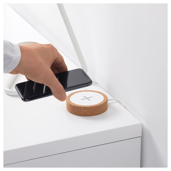 A person places a smartphone on a round wireless charger made of cork. The charger sits on a white surface.