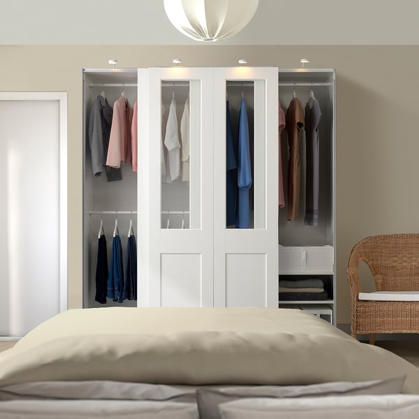 Modern bedroom with white PAX wardrobe, glass panels, and sliding doors. Beige bed, light brown pillow, wicker chair with cushion.