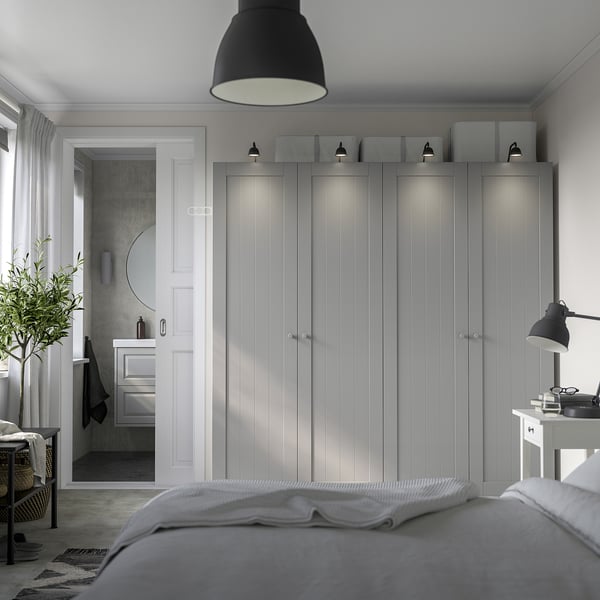 Minimalist bedroom with large grey PAX wardrobe, integrated lighting, desk lamp, cosy bed, and small bathroom with round mirror.