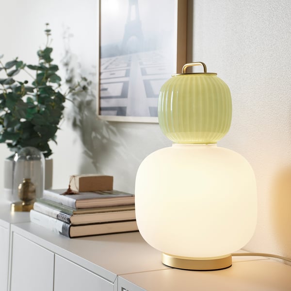 Modern, yellow-green & white PILBLIXT lamp with glass & metal, frosted finish, sits on shelf beside books & plant, small.