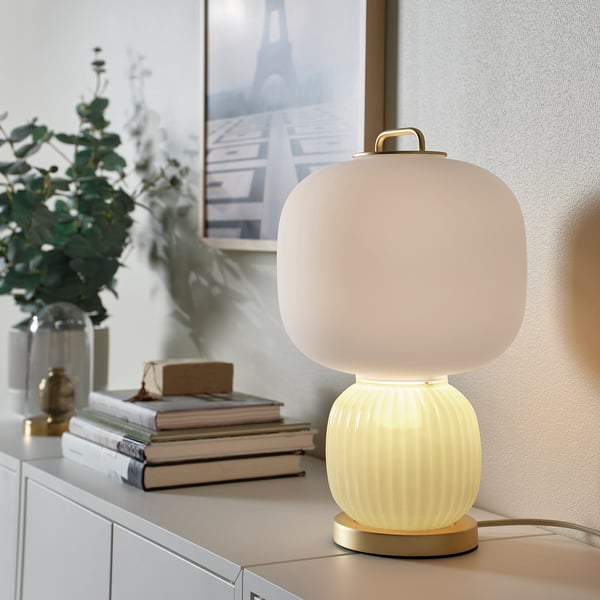 A modern PILBLIXT lamp with a frosted glass shade on a metal base. It stands next to books and a plant on a cabinet, emphasising its compact size.