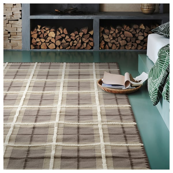 A brown and grey wool rug with a classic tartan check pattern, displayed in a room with a green bench and stacked firewood.