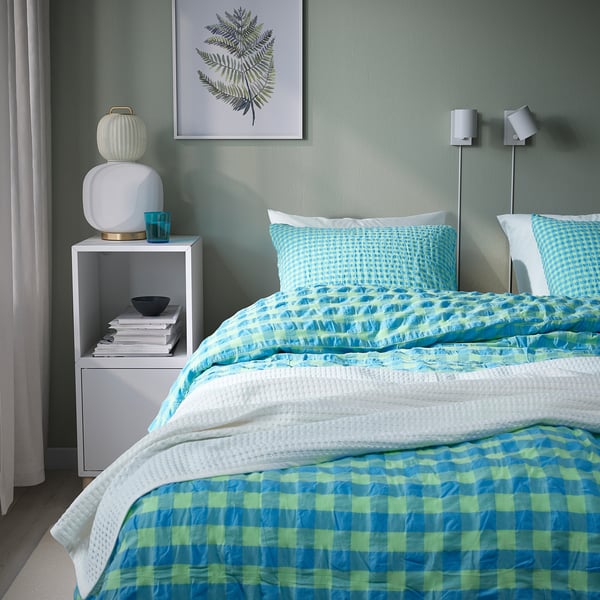 Bed with turquoise & lime RÅGBLOMMA duvet, cotton-viscose, large, pizza-shaped pillows, white blanket. Beside, lamp & books on small shelf.