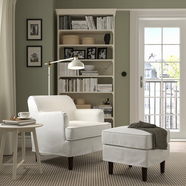 Cosy living room with ROCKSJÖN armchair, footstool, and book-filled shelf. Green walls, natural light, and a sweater-draped footstool.