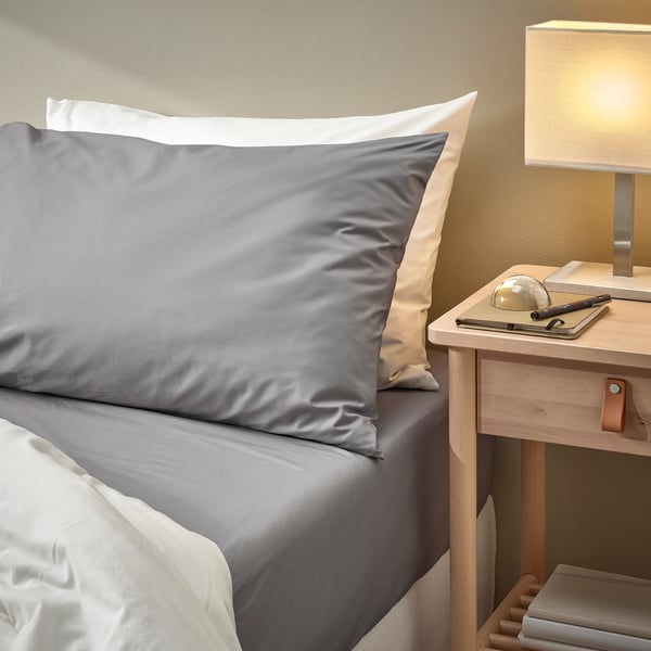 Nearby a bed with grey RÖNNVECKMAL sheets, a wooden bedside table holds a lamp, books, and a globe. The pillowcases neatly frame the headrest.