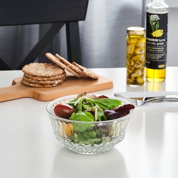Clear glass bowl filled with salad sits on a white table, accompanied by crackers and condiments.