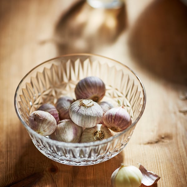 Clear glass bowl filled with garlic bulbs, elegantly patterned, on a wood table.