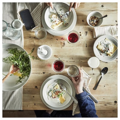 A rustic dinner setting with wooden table, SANDSKÄDDA stoneware plates, bowls, glasses, and cutlery. People are enjoying food and drinks.