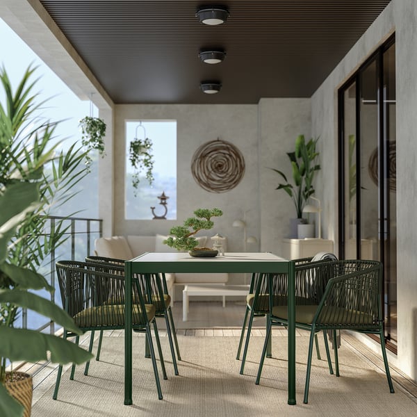 Modern outdoor dining set: rectangular green metal table with woven chairs, surrounded by plants on a balcony.