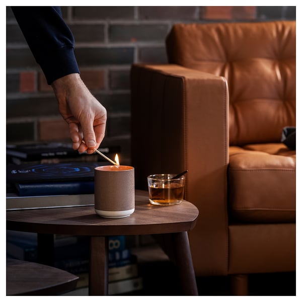 Hand lights silk candle on wooden table next to amber drink on leather couch, cosy brick background.