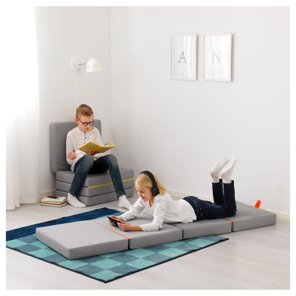 Two kids relax in a minimalist room. One reads on a grey modular mattress, another scrolls on a smartphone while lying on another folded mattress.