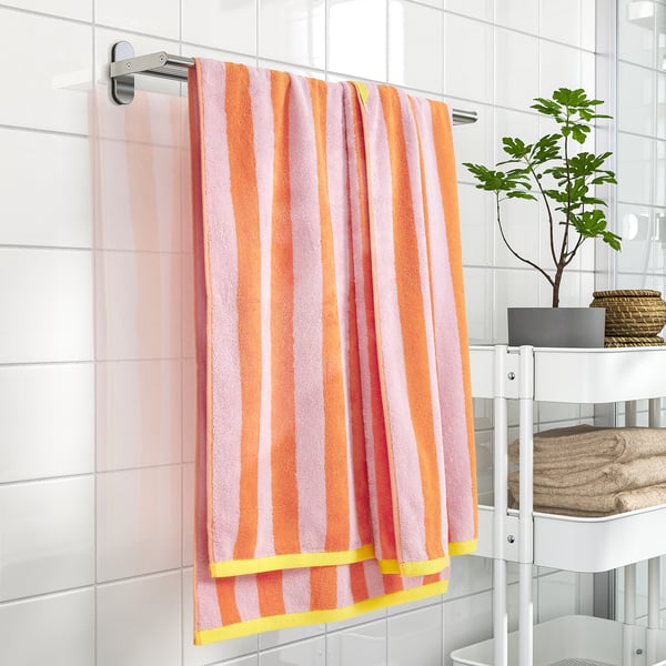 Striped SLÅNHÖSTMAL towel hangs on wall rack, bright colours, 100% cotton, framed by tiles, next to small plant and folded towels on shelf.