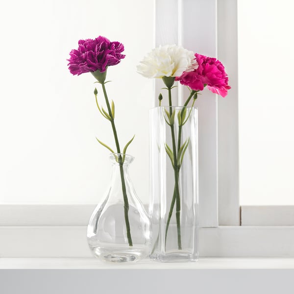 Two clear glass vases hold colourful artificial flowers on a windowsill. One vase contains purple flowers, the other white and pink.