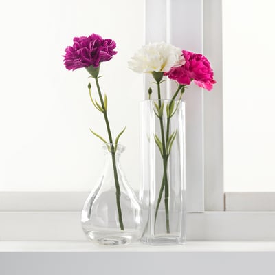 Two clear glass vases hold colourful artificial flowers on a windowsill. One vase contains purple flowers, the other white and pink.