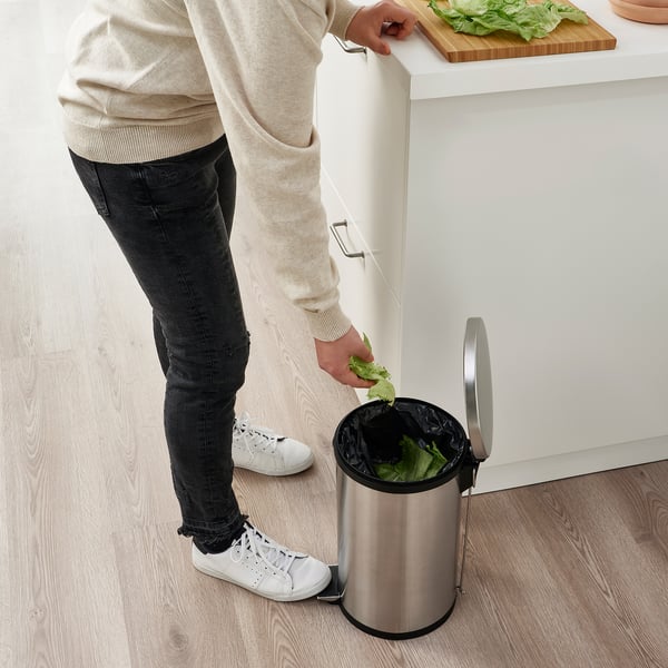 Person disposing of lettuce into a stainless steel pedal bin with a handle on the back, positioned under a countertop.