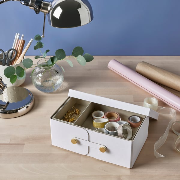 White storage box with two compartments and gold knobs holds mbc. Next to lamp, pens, and wrapping paper on wood desk.