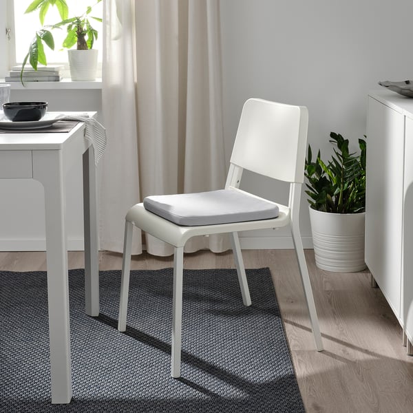 Minimalist dining area: white staggastrarr chair, grey cushion, near table & plant. Light-filled.