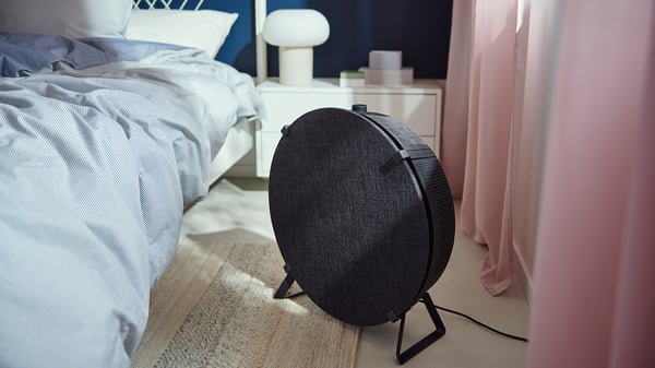 In a cosy bedroom, a black cylindrical IKEA STARKVIND air purifier stands on a rug beside a bed with blue and white linens and a pink curtain.