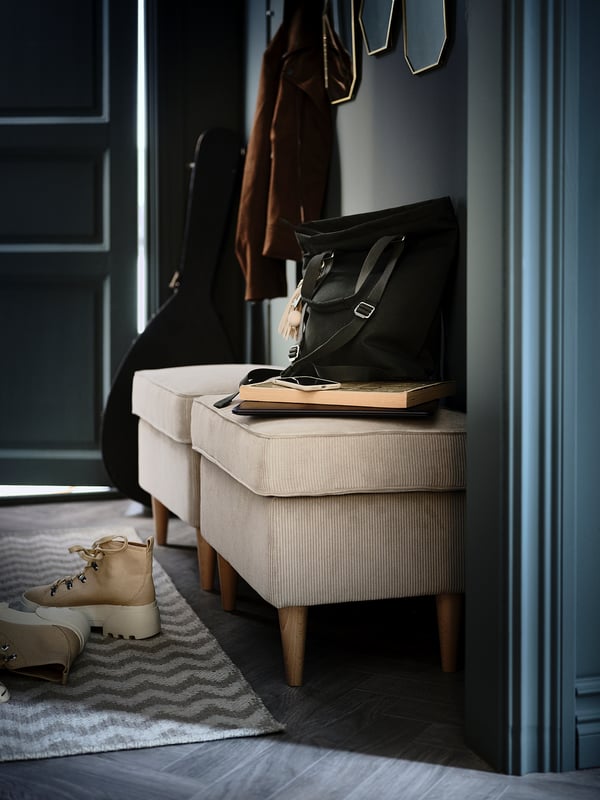 Cosy entryway scene with light beige STRANDMON footstool, black bag, books, and tan boots on zigzag rug under coat rack.
