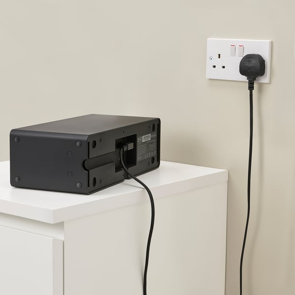 Black speaker on white table, plugged into wall socket with long cord.