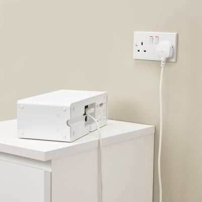 White speaker on white shelf, plugged into wall socket with long cord.