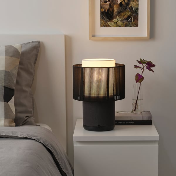 A bed with a SYMFONISK lamp on a bedside table, featuring a black base, fabric shade, flower vase, and pillow.