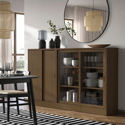Wooden TONSTAD sideboard with glass doors, mirror, dishes, wicker chair, rug, pendant lamps, minimalist decor.