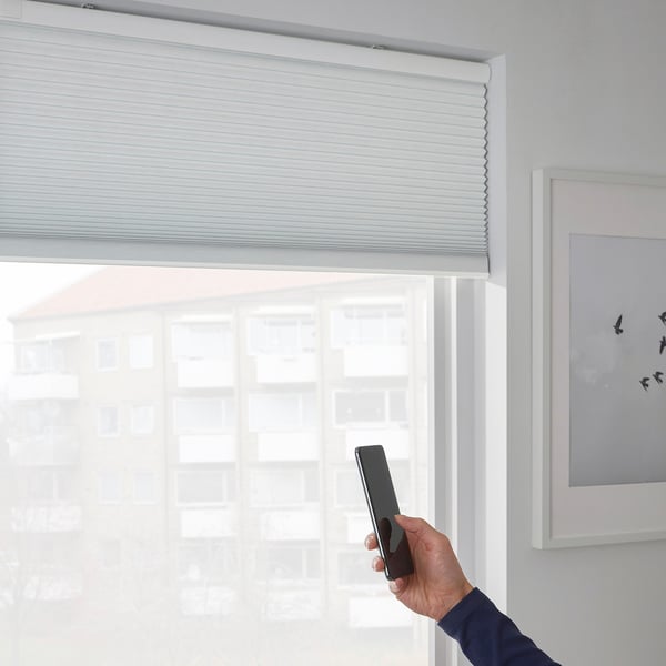 Hand holding phone adjusts smart blinds via app. Blinds cover window, blocking light and draughts.