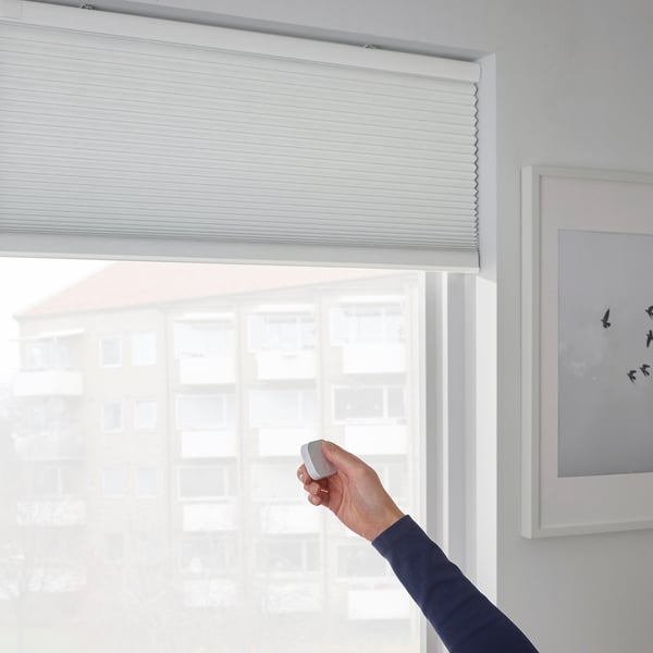 A hand raises a remote control adjusting a cordless TREDANSEN cellular blind. The blind philtres light and insulates, enhancing privacy and comfort.