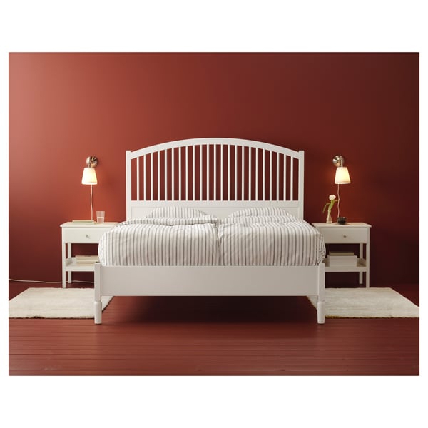 A minimalist white wooden bed with striped bedding sits against a reddish wall. Two nightstands with lamps flank it.