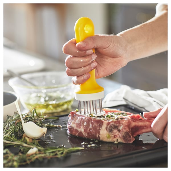 A chef uses a yellow UPPFYLLD meat tenderizer with metal spikes on a steak surrounded by herbs and garlic, enhancing flavour.