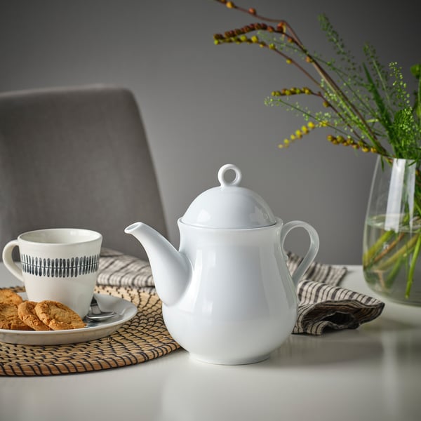 White porcelain teapot with lid and handle sits on woven mat beside a mug with cookies and a vase with flowers.