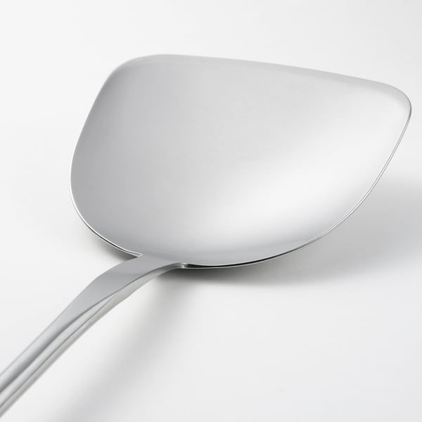 A sleek, stainless steel wok spatula with a long handle, resting on a plain white surface, showcasing its polished, well-crafted design.