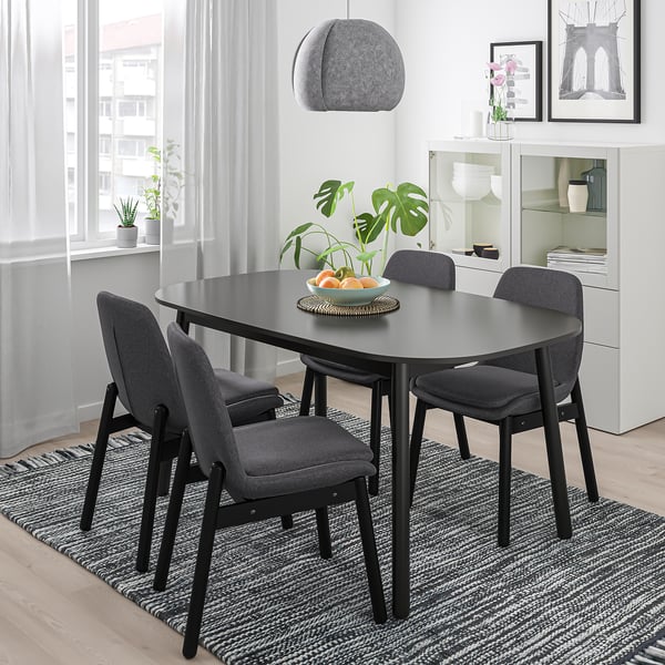 Modern dining room with black oval VEDBO table, four grey chairs, pendant lamp, plant, and fruit bowl.