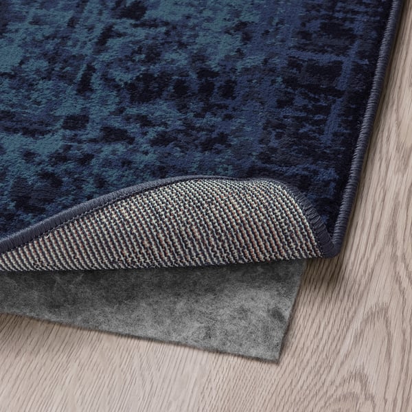 Dark blue patterned rug with raised edge detail, placed on light wooden floor, showcasing its low pile and non-slip padding.