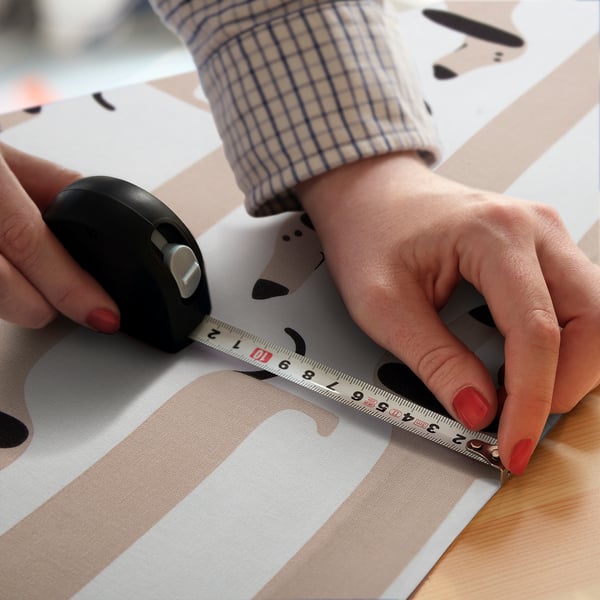 A person measures abetstag fabric, marked with playful dachshunds, using a tape and a mouse on a white desk.