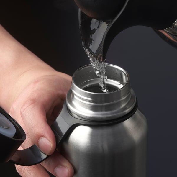 Person pouring water into a shiny, insulated, stainless steel thermos flask. The thermos has a secure lid and is held in one hand.