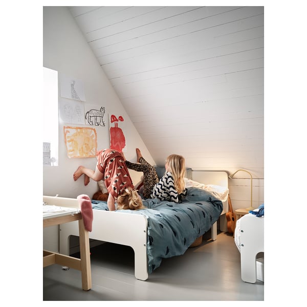 Two children playing on a bed with a blue BARNDRÖM duvet cover, featuring white car prints. The room has slanted white walls and colourful artwork.