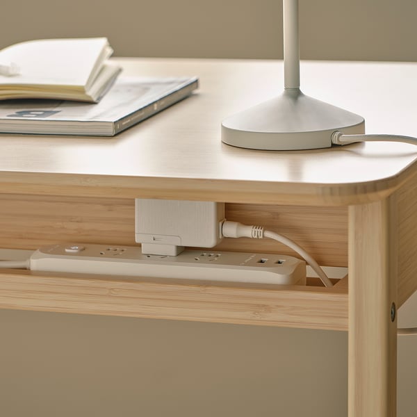 A wooden desk with cable management and a drawer. A lamp and books are on top.