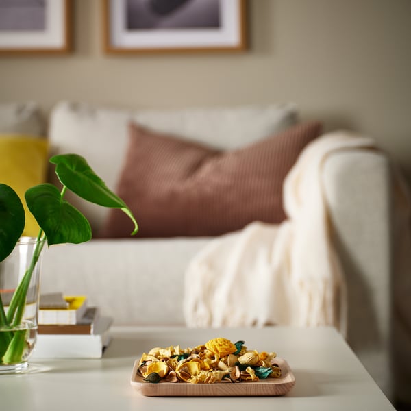 Wooden BROKFINK dish holds potpourri on living room table, near white sofa with pillows, beside a plant in a vase.