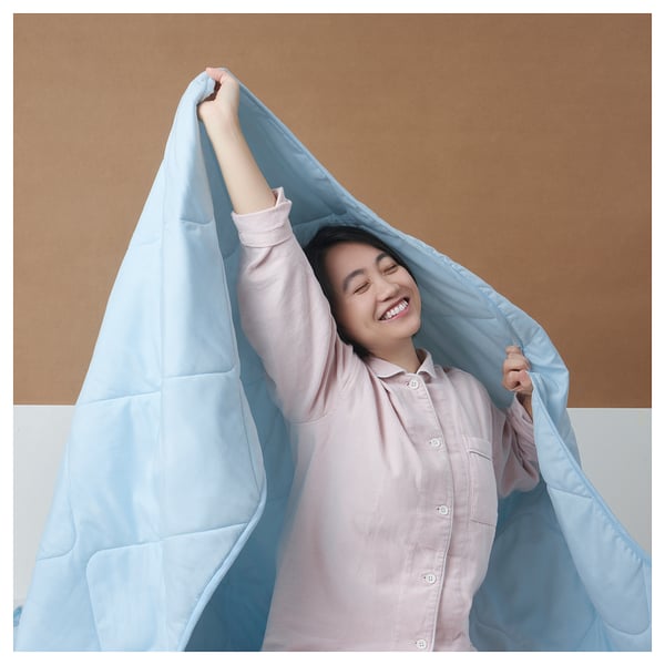A person holding a light blue BRUKSVARA duvet, smiling, over a brown background.