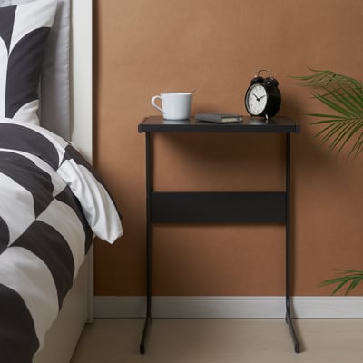 Small black side table next to bed, holding a white mug, alarm clock, and magazines.