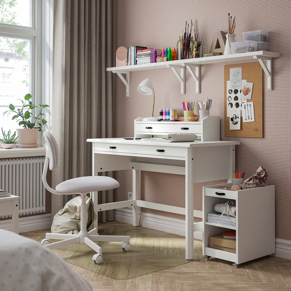 White BRYTNING desk, organised with storage, positioned beside a window with curtains. Its compact size fits neatly in the corner.