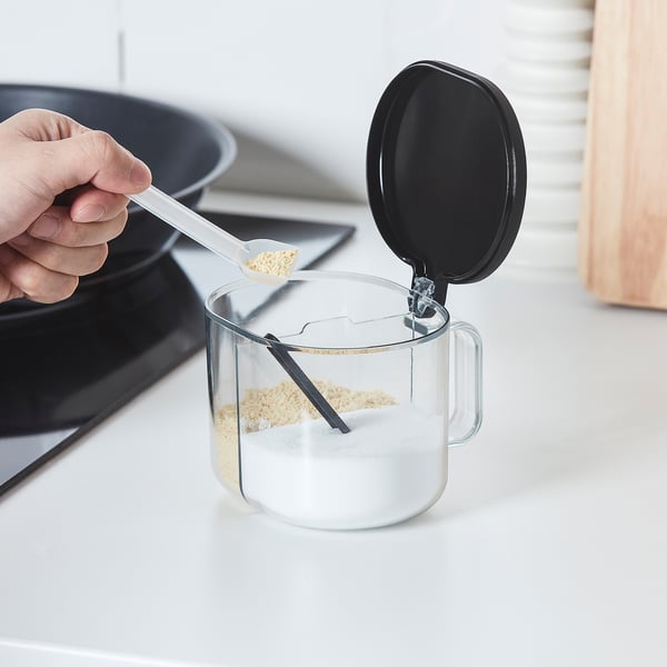 Hand pours seasoning from spoon into clear, two-compartment jar with opening lid, ideal for kitchen convenience.
